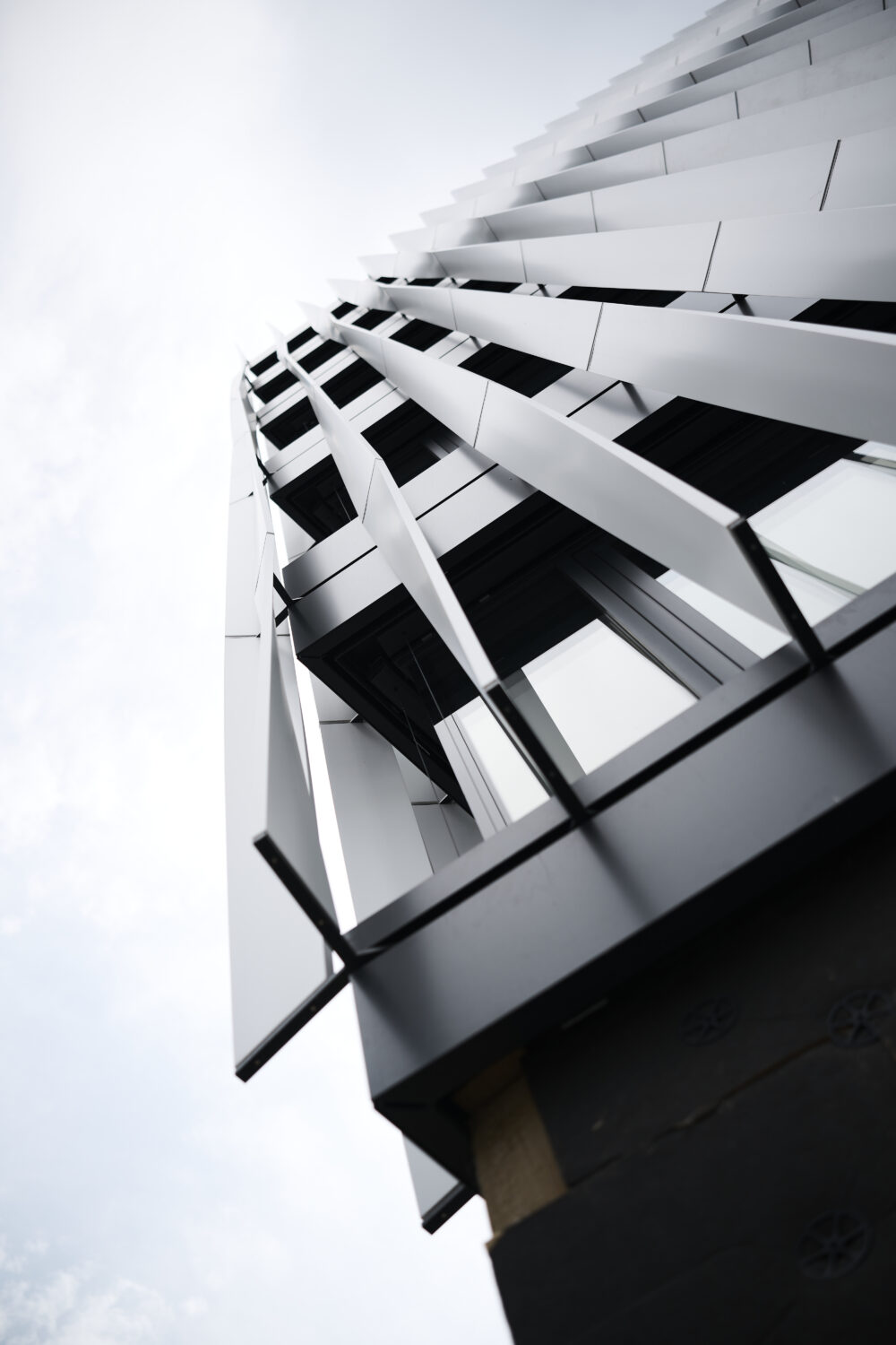 Eine moderne Gebäudefassade mit schräg angeordneten Metallpaneelen und großen Fenstern, von unten vor einem bewölkten Himmel betrachtet.