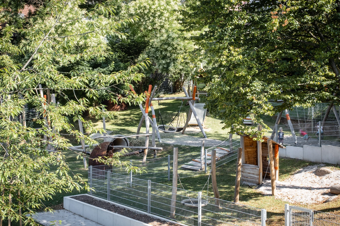 Ein umzäunter Spielplatz mit Schaukeln, einem Tunnel und einem kleinen Holzunterstand – der in der Projektentwicklungsphase von SCHATZ Architektur fachmännisch entworfen wurde – ist an einem sonnigen Tag von Bäumen und Grünflächen umgeben.