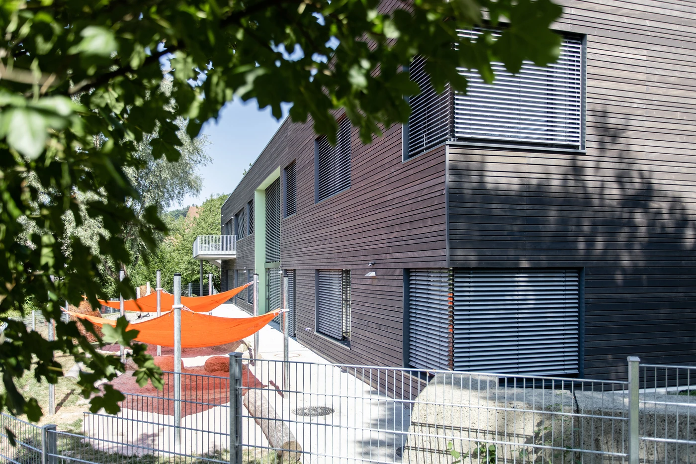 Ein modernes, von SCHATZ Architektur entworfenes Gebäude mit horizontaler Holzverkleidung und großen Fenstern wird teilweise von üppigen Ästen beschattet. Orangefarbene Sonnenschirme überdachen einen umzäunten Spielplatz im Außenbereich. Die Szene spiegelt die helle und sonnige Atmosphäre wider, die durch eine durchdachte Projektentwicklung und fachkundig gesteuerte Leistungsphasen erreicht wurde.
