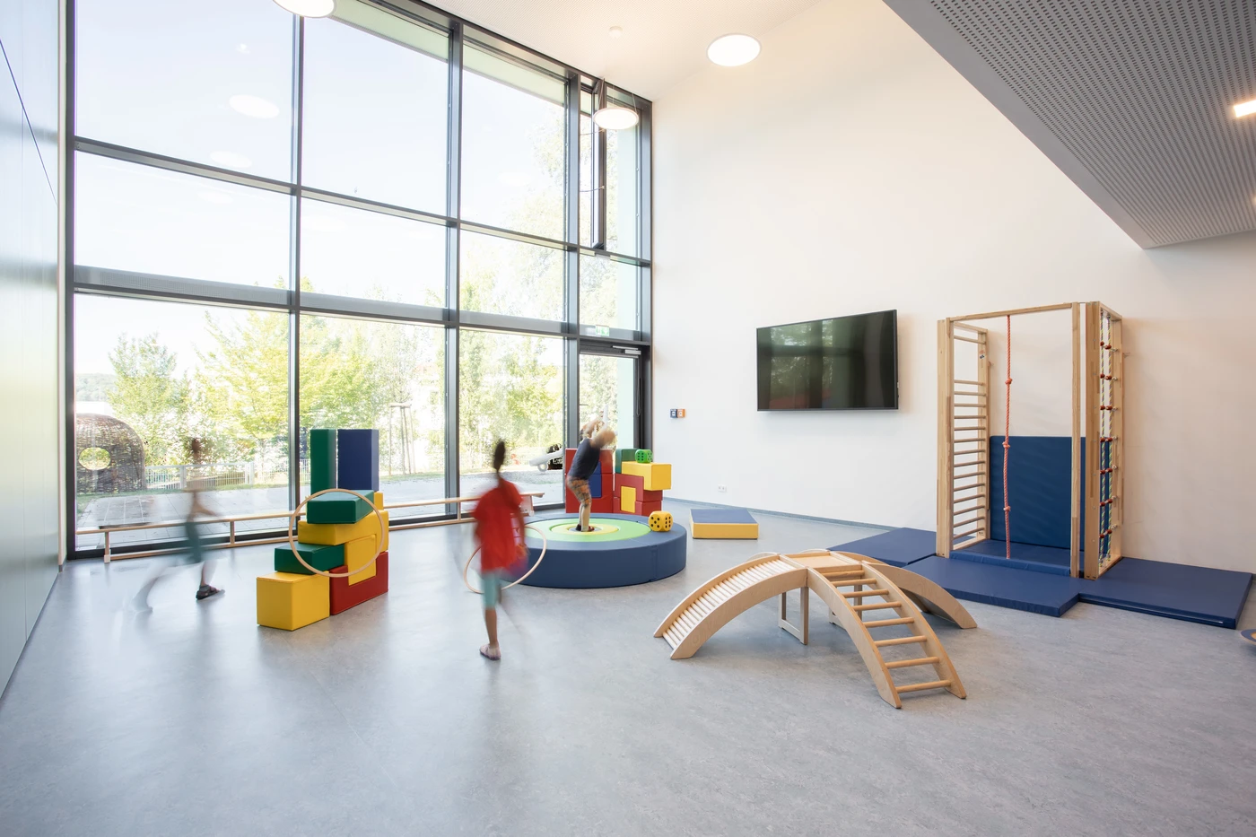 Die Kinder spielen in einem hellen Raum mit großen Fenstern, weichen Spielklötzen, einer Kletterwand, einer Rutsche und einer Holzbrücke – einem einladenden Raum, der von SCHATZ Architektur in der Entwurfsphase sorgfältig gestaltet wurde. Das natürliche Licht und die minimalistische Einrichtung des Raums schaffen eine Atmosphäre, die zum Entdecken und zur Kreativität anregt.