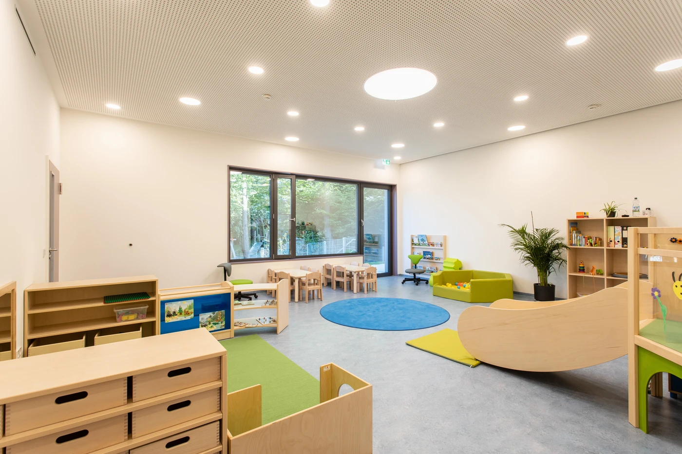 Ein helles, modernes Vorschulzimmer, das von SCHATZ Architektur in Schorndorf entworfen wurde, verfügt über Holzmöbel, einen blauen Teppich, grüne Sitzgelegenheiten, Bücherregale, Spielzeug, Pflanzen und große Fenster, durch die Tageslicht hereinströmt.