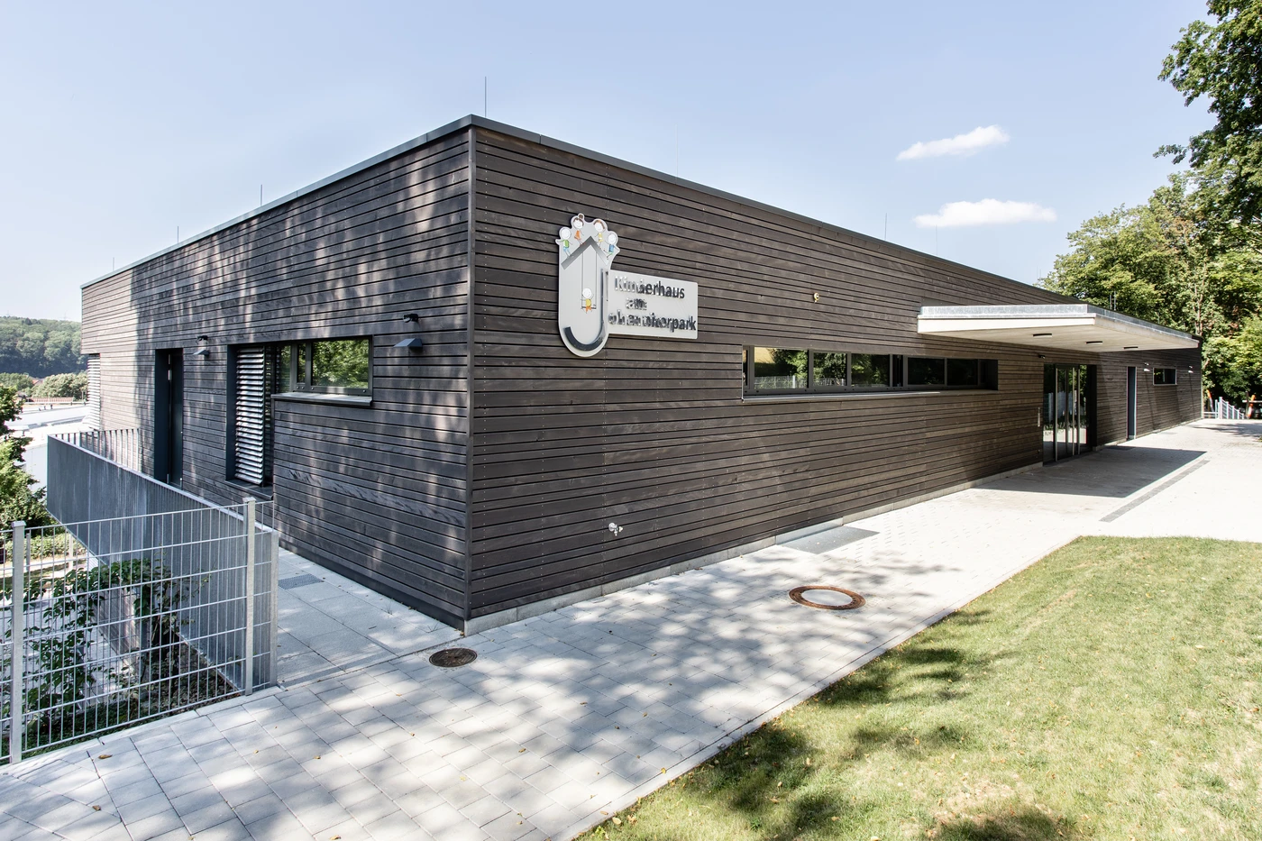 Bildung Ein modernes, rechteckiges Gebäude mit dunkler Holzverkleidung, großen Fenstern und einem Flachdach, umgeben von Rasen, Bäumen und einem gepflasterten Gehweg. Entworfen von SCHATZ Architektur im Rahmen ihrer Generalplanung für Schorndorf; in der Nähe des Eingangs ist ein schildförmiges Schild angebracht.