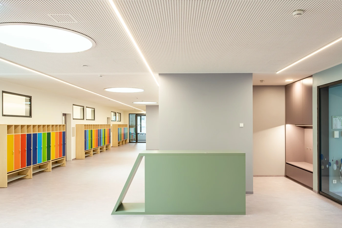 Ein moderner Schulflur, entworfen von SCHATZ Architektur, besticht durch farbenfrohe Spinde auf der linken Seite, einen grünen Empfangstresen in der Mitte und graue Sitzbereiche auf der rechten Seite unter hellen Deckenleuchten – ein Beispiel für die Kompetenz des Büros in den Bereichen Generalplanung und allen Leistungsphasen.