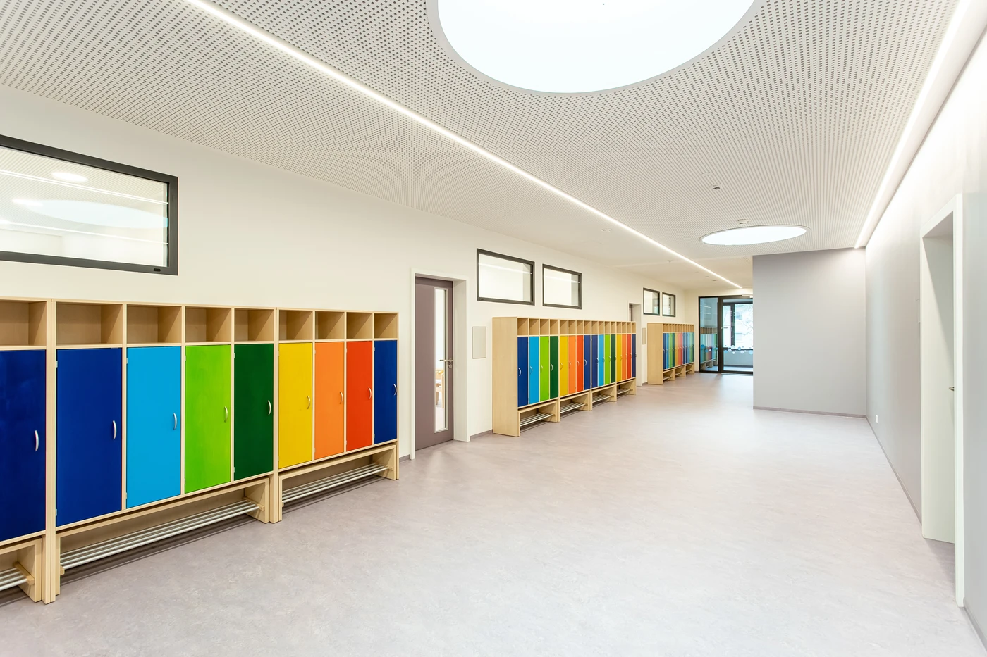 Ein heller Schulflur mit bunten Spinden in Blau, Grün, Orange und Gelb, hellen Wänden, großen Fenstern und einer modernen Decke mit runden Leuchten zeugt von einer durchdachten Planung und Projektentwicklung im Rahmen der Gesamtplanung.