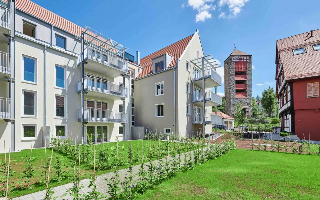 Projekte Moderne Wohnanlagen mit Balkonen und einem gepflegten grünen Rasen im Vordergrund, die die für SCHATZ Architektur charakteristische Liebe zum Detail widerspiegeln. Im Hintergrund sind unter einem strahlend blauen Himmel ein Steinturm und ein weiteres Gebäude zu sehen, was die durchdachte Gesamtplanung in jedem Aspekt dieser Projektentwicklung verdeutlicht.