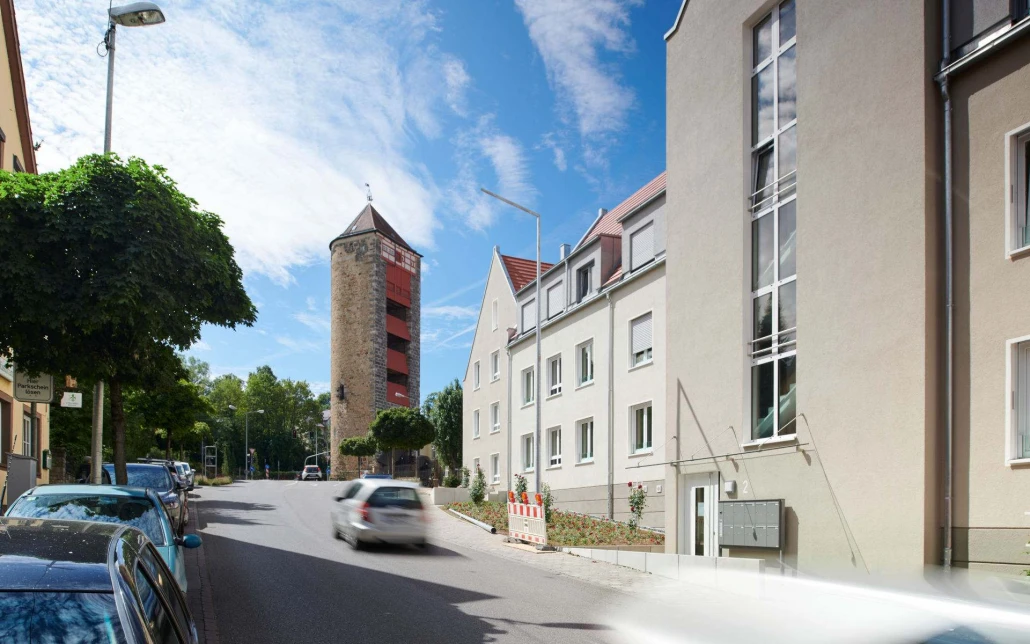 Projekte Ein Auto fährt eine sonnige Straße entlang, die von geparkten Fahrzeugen und modernen Gebäuden gesäumt ist – ein Zeugnis durchdachter Projektentwicklung; am Ende der Straße erhebt sich unter einem blauen Himmel mit vereinzelten Wolken ein alter Steinturm, der dem Entwurf von SCHATZ Architektur für dieses Gebiet historischen Charme verleiht.
