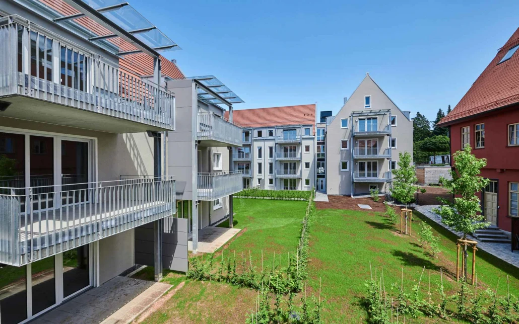 Projekte Moderne Mehrfamilienhäuser mit Balkonen, die durch einen durchdachten Entwurf geprägt sind, umgeben einen begrünten Innenhof in Schorndorf. Die Gebäude zeichnen sich durch helle Fassaden und rotgedeckte Dächer aus, ergänzt durch kleine, neu gepflanzte Bäume und einen strahlend blauen Himmel – ein Paradebeispiel für zeitgenössische Generalplanung.