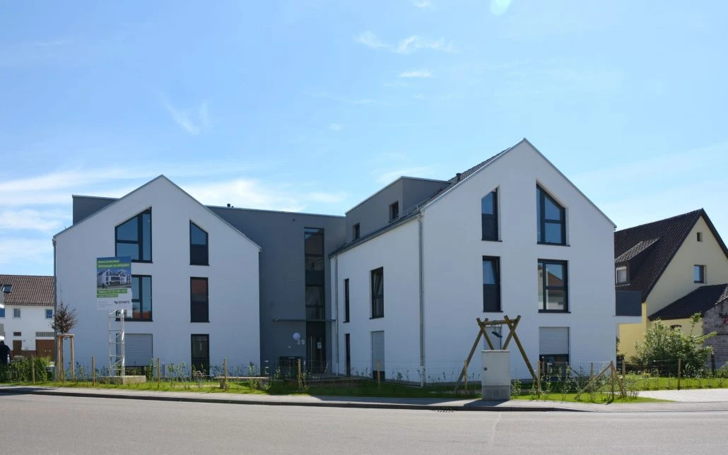 Modernes weißes Wohnhaus mit markanten Dachwinkeln, großen Fenstern und einem kleinen Vorgarten, aufgenommen an einem sonnigen Tag mit strahlend blauem Himmel. Als Beispiel für durchdachte Projektentwicklung und Generalplanung bietet diese Immobilie zeitgemäßes Wohnen im Herzen von Schorndorf.