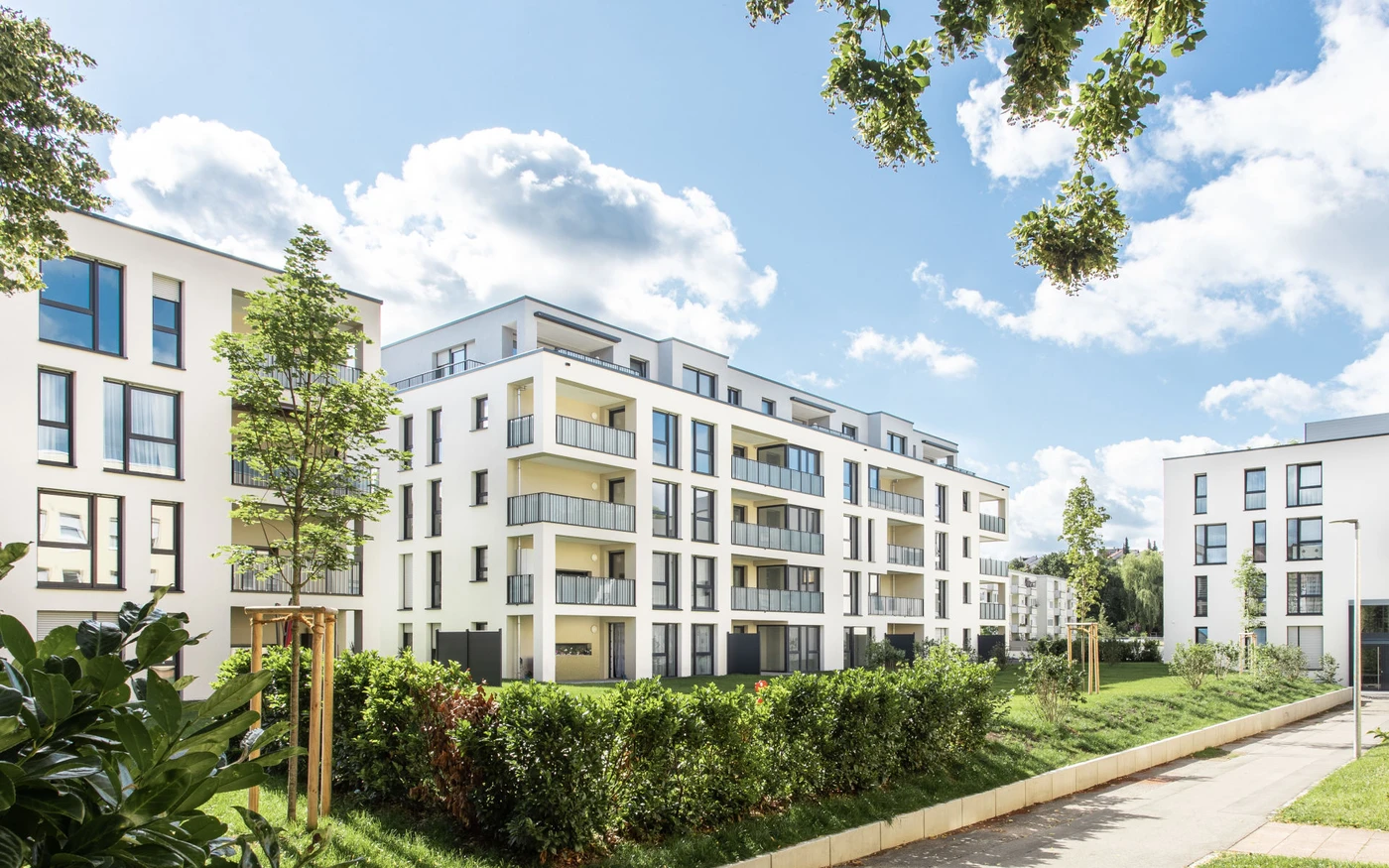 Moderne weiße Mehrfamilienhäuser mit Balkonen, großen Fenstern und begrünten Außenanlagen unter einem teilweise bewölkten Himmel in Schorndorf. Bäume und Sträucher säumen den Weg vor den Gebäuden und zeugen von einer durchdachten Gesamtplanung über alle Leistungsphasen hinweg.