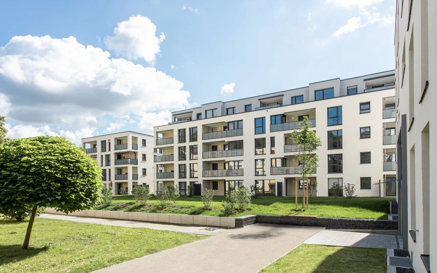 Moderne Wohnanlagen mit Balkonen und großen Fenstern, die durch sorgfältige Entwurfs- und Generalplanung durchdacht gestaltet wurden, sind umgeben von grünen Rasenflächen, jungen Bäumen und einem blauen Himmel mit vereinzelten Wolken. Ein gepflasterter Weg führt durch die landschaftlich gestaltete Anlage und spiegelt auf Schritt und Tritt die sorgfältige Projektentwicklung wider.