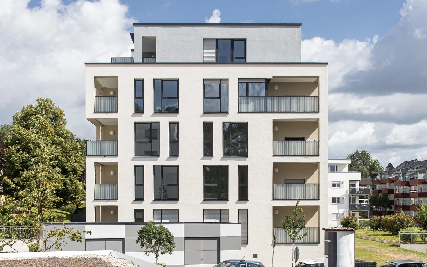 Ein modernes, weißes Mehrfamilienhaus mit zahlreichen Balkonen und großen Fenstern, umgeben von Grünflächen und geparkten Autos, an einem teilweise bewölkten Tag – ein Beispiel für den innovativen Ansatz von SCHATZ Architektur in den Bereichen Projektentwicklung und Generalplanung.