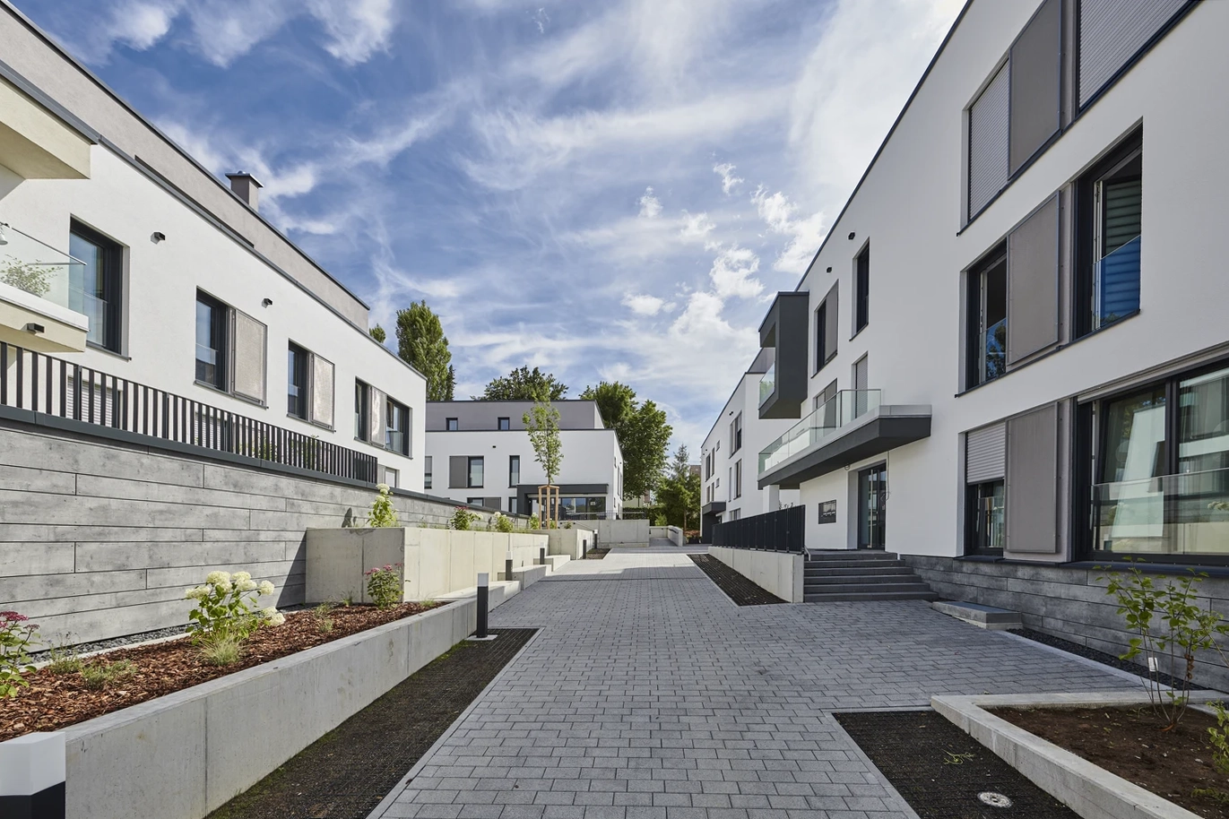 Projekte Moderne Wohnblocks mit weißen Fassaden und großen Fenstern, die durch einen breiten, gepflasterten Fußweg im Herzen von Schorndorf voneinander getrennt sind. Hochbeete säumen den Weg und zeugen unter teilweise bewölktem Himmel von einem durchdachten Entwurf und viel Liebe zum Detail.