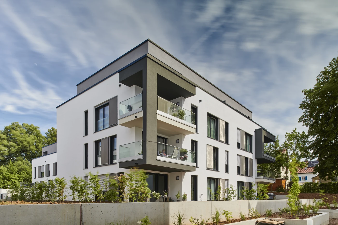 Projekte Modernes weißes Mehrfamilienhaus mit großen Fenstern, Glasbalkonen und einer begrünten Außenanlage vor dem Gebäude. Diese beeindruckende Anlage in Schorndorf ist ein Paradebeispiel für durchdachte Projektentwicklung mit Schwerpunkt auf hochwertiger Generalplanung. Der Himmel ist teilweise bewölkt, und das Gebäude ist von Bäumen umgeben.