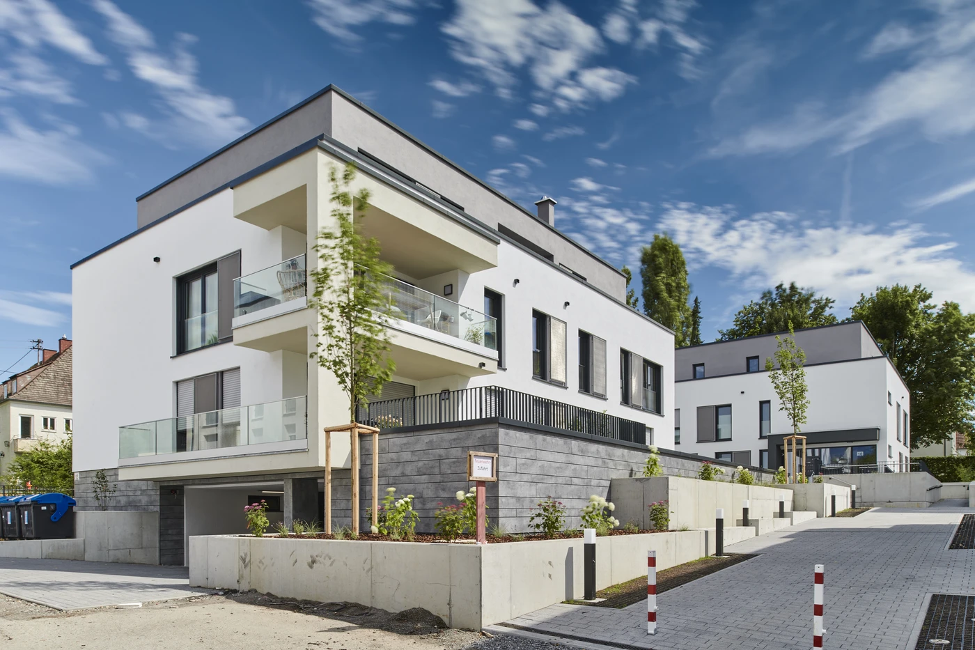 Projekte Modernes weißes Mehrfamilienhaus mit großen Balkonen, Glasgeländern und einer gepflegten Außenanlage unter blauem Himmel mit vereinzelten Wolken in Schorndorf. Im Hintergrund ist ein weiteres, ähnliches Gebäude zu sehen, was die sorgfältige Projektentwicklung über alle Leistungsphasen hinweg widerspiegelt.