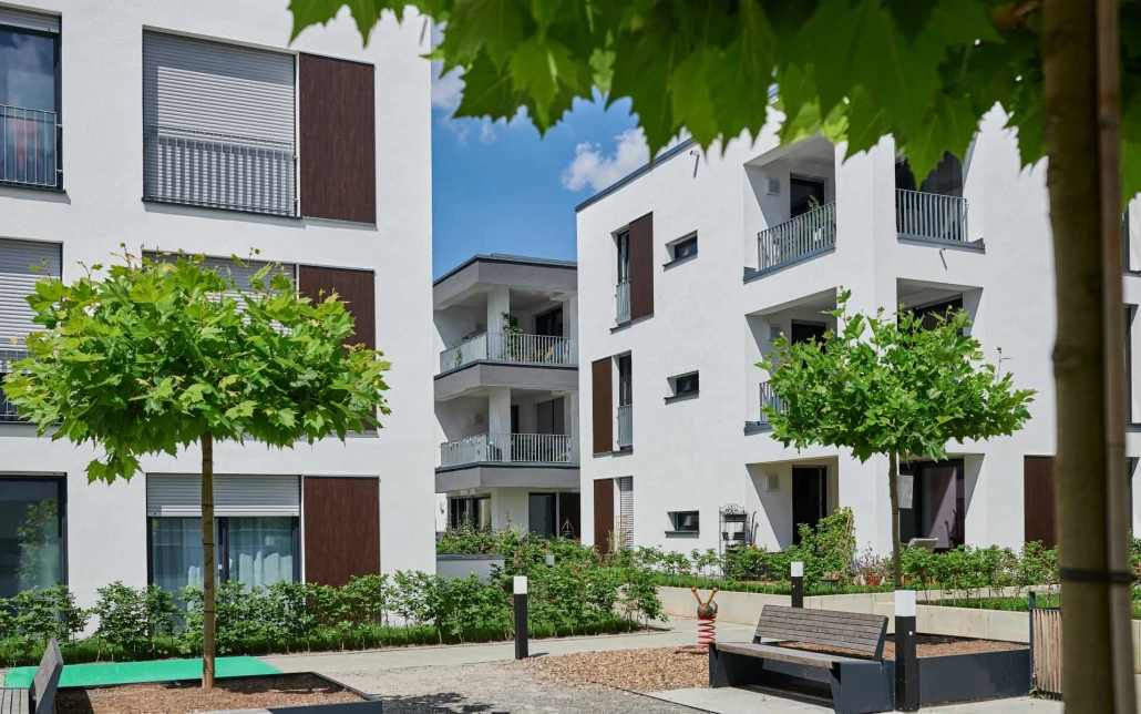 Projekte Moderne weiße Mehrfamilienhäuser mit Balkonen umgeben einen kleinen Innenhof mit Bänken, jungen Bäumen, Pflanzen und einem strahlend blauen Himmel – ein durchdachter Entwurf, der vom zeitgenössischen Wohnen in Schorndorf inspiriert ist und über alle relevanten Leistungsphasen hinweg entwickelt wurde.