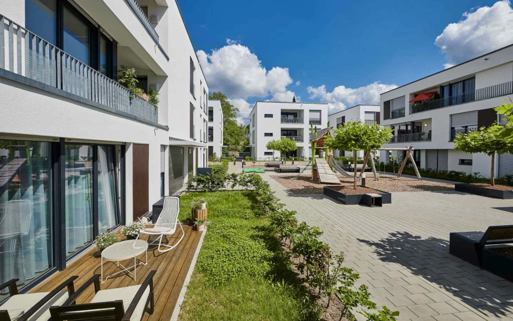 Projekte Moderne Mehrfamilienhäuser mit Balkonen und Terrassen, die im Rahmen eines Entwurfs von SCHATZ Architektur entstanden sind, umgeben einen begrünten Innenhof mit Bänken, jungen Bäumen, einem Spielplatz mit Rutsche und einer Schaukel unter blauem Himmel mit flauschigen Wolken.