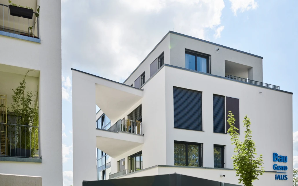 Projekte Ein modernes, weißes Mehrfamilienhaus mit großen Fenstern, Balkonen und schwarzen Fensterläden vor einem teilweise bewölkten Himmel. Das Gebäude, das im Rahmen eines innovativen Entwurfs in Schorndorf entstanden ist, wird im Vordergrund von einem kleinen Baum und einem Teil eines anderen Gebäudes eingerahmt – ein Beispiel für durchdachte Projektentwicklung bis ins kleinste Detail.