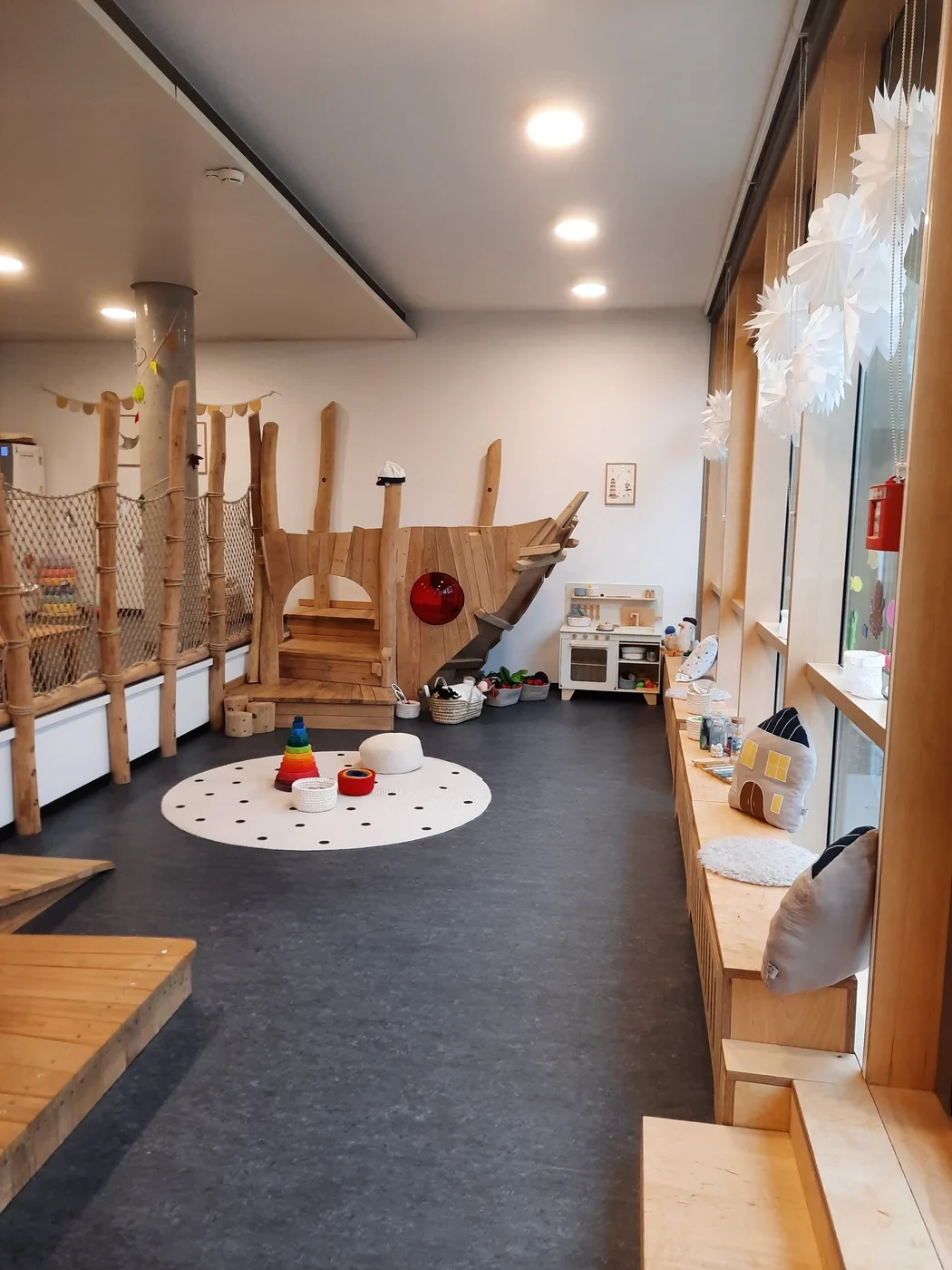 Bildung Ein gemütliches, helles Spielzimmer – das durch durchdachten Entwurf und Generalplanung sorgfältig gestaltet wurde – verfügt über Spielgeräte aus Holz, eine Rutsche, einen weißen Teppich mit Polka-Punkten und Spielzeug sowie große Fenster, die mit Papierschneeflocken geschmückt sind. Kissen säumen die Fensterbank und spiegeln die Liebe zum Detail in allen Leistungsphasen wider.