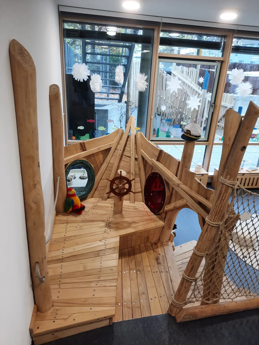 Holzspielgerüst für den Innenbereich in Form eines Schiffes, mit Steuerrad, Bullaugen, einer Seilbrücke und einem Plüschpapagei. Große Fenster im Hintergrund geben den Blick auf einen mit Schneeflocken geschmückten Spielbereich im Freien frei – ein einladender Ort, der die durchdachte Projektentwicklung und kreative Planung in Schorndorf unterstreicht.