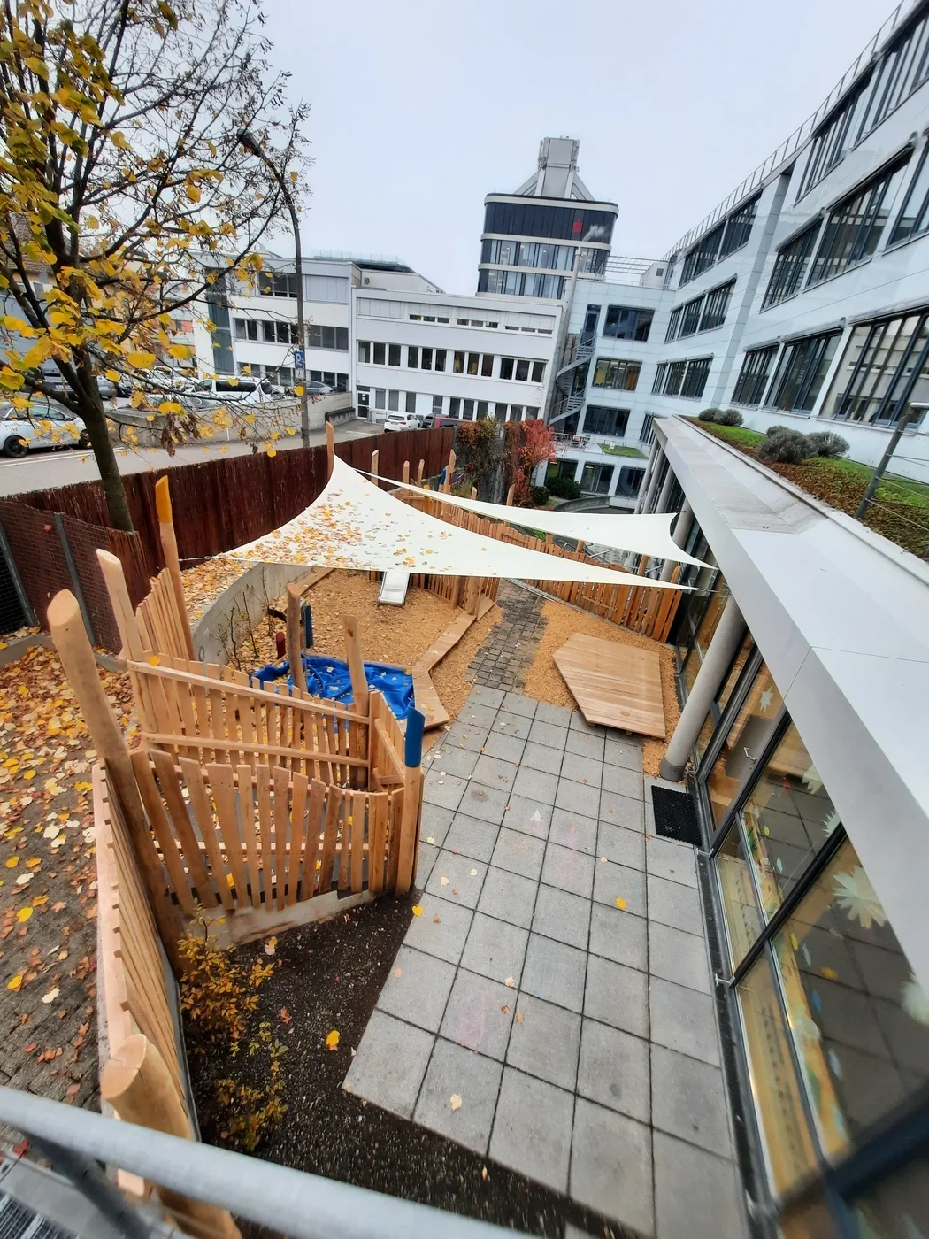 Ein eingezäunter Spielplatz im Freien mit einem Sonnensegel, einem Sandkasten, Holzkonstruktionen und herabgefallenem Herbstlaub, der im Rahmen einer Projektentwicklung in Schorndorf gestaltet wurde, ist an einem bewölkten Tag von modernen Bürogebäuden umgeben.