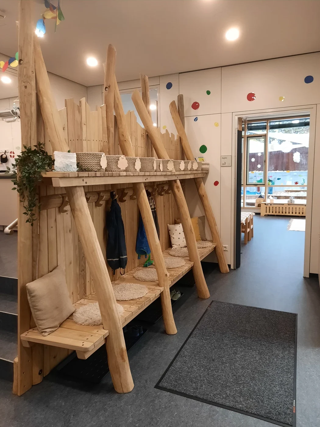 Eine gemütliche Garderobe in einer Vorschule in Schorndorf mit Holzbänken, Kleiderhaken, Kissen, Pflanzen und bunten Punkt-Aufklebern an den Wänden zeugt von einer durchdachten Projektentwicklung. Durch eine offene Tür ist ein Klassenzimmer mit niedrigen Tischen zu sehen.