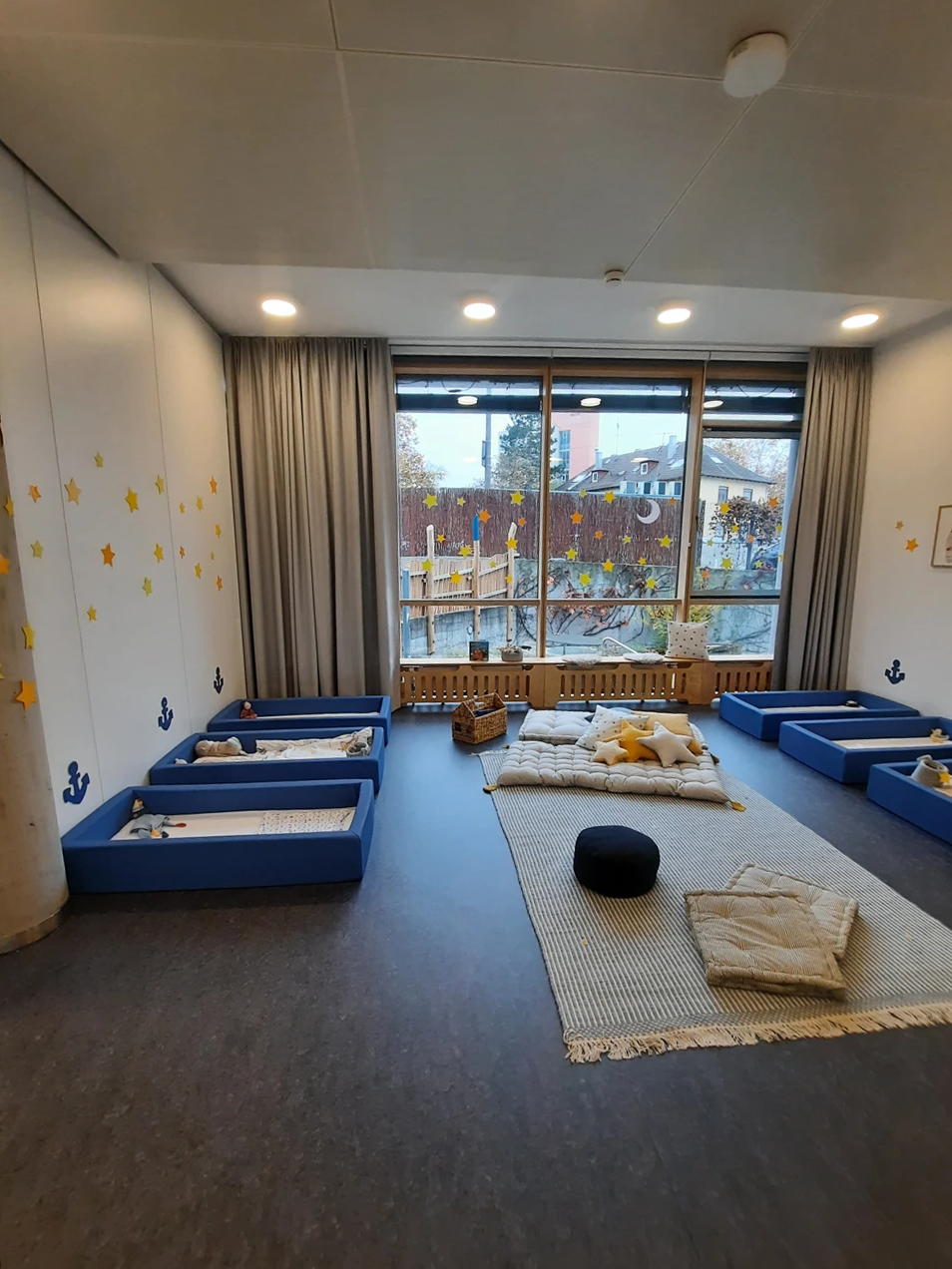 Ein gemütlicher Mittagsschlafraum in einer Kindertagesstätte, entworfen von SCHATZ Architektur, mit blauen Kleinkinderbetten, einem Teppich mit Kissen und Decken, gelben Stern-Wandaufklebern und großen Fenstern mit Blick auf einen Spielplatz in Schorndorf.