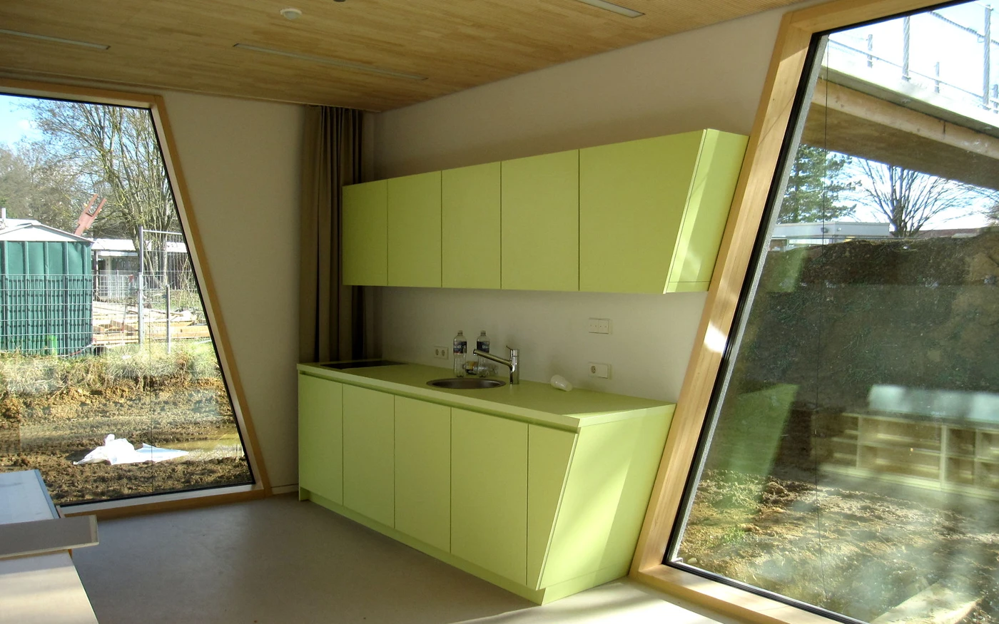 Eine moderne Küche mit hellgrünen Schränken, einer Spüle und großen, schräg gestellten Fenstern, die Tageslicht hereinlassen und den Blick auf eine Baustelle im Freien freigeben – die perfekte Inspiration während der Entwurfsphase jeder Projektentwicklung.