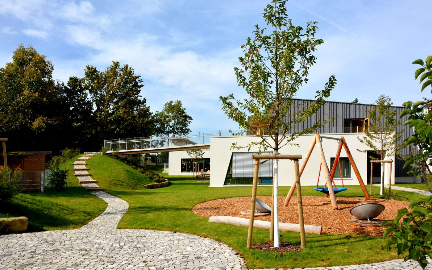 Bildung Ein moderner Spielplatz mit Schaukeln, Klettergerüsten, einem geschwungenen Steinweg, grünem Rasen, jungen Bäumen und einem zeitgenössischen Gebäude im Hintergrund unter blauem Himmel – ein einladender Freiraum, der durch sorgfältige Entwurf und Generalplanung in Schorndorf gestaltet wurde.
