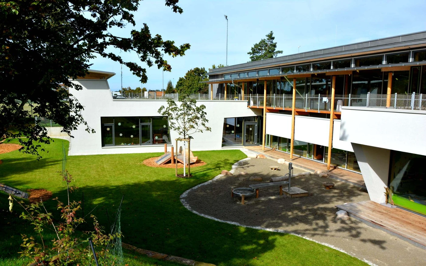 Ein modernes Schulgebäude mit großen Fenstern, entworfen von SCHATZ Architektur, umgibt einen begrünten Innenhof mit einem kleinen Spielplatz, einem Sandkasten und Bäumen unter freiem Himmel. Das Projekt steht beispielhaft für eine umfassende Generalplanung über alle Leistungsphasen hinweg.
