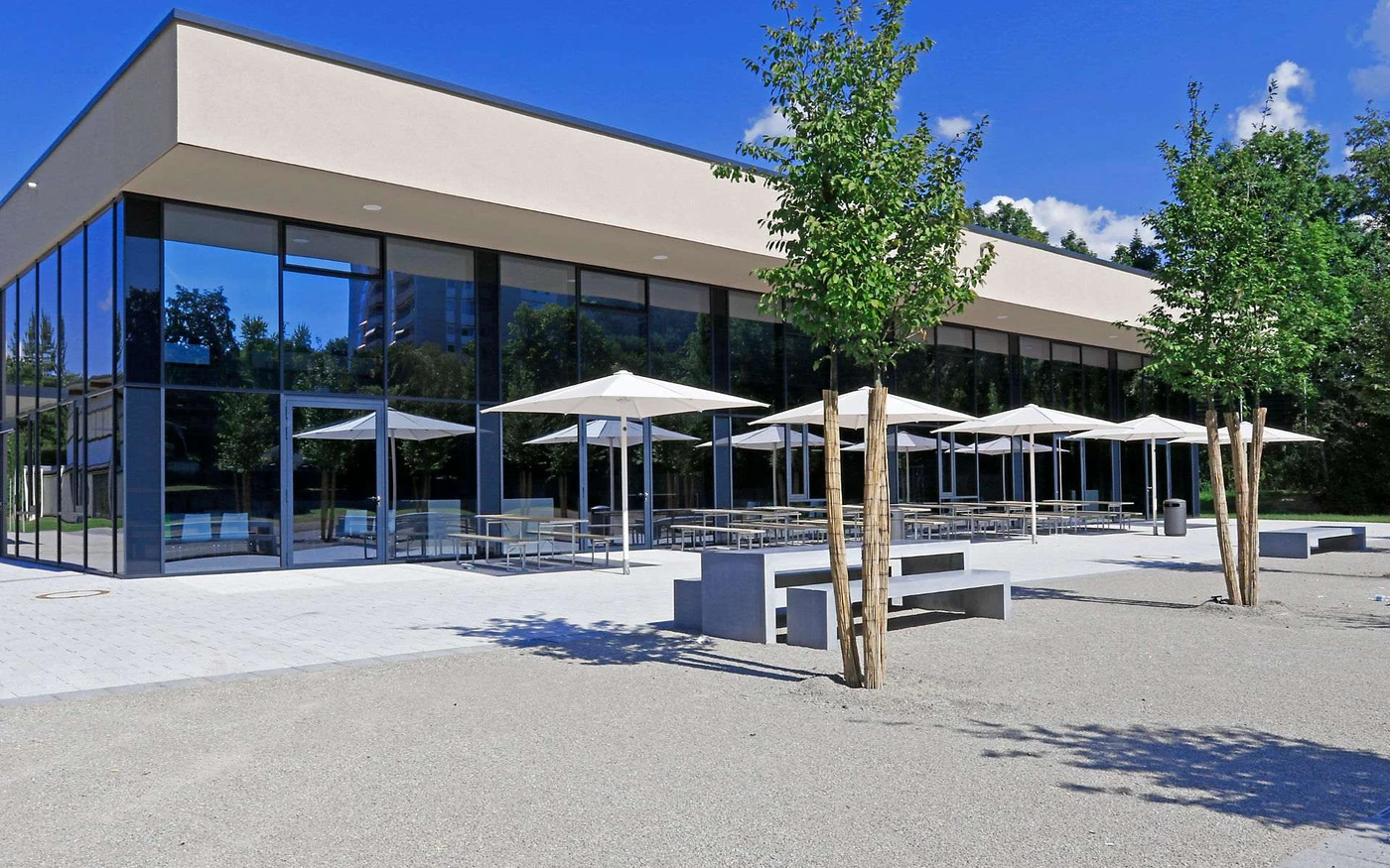 Bildung Ein modernes Gebäude mit großen Glasfenstern, Sitzgelegenheiten im Freien unter weißen Sonnenschirmen und mehreren Bäumen, die auf einem Kieshof unter strahlend blauem Himmel gepflanzt sind – ein inspirierendes Beispiel für zeitgenössischen Entwurf und durchdachte Gesamtplanung.
