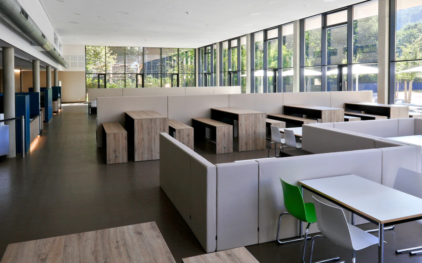 Moderne Cafeteria mit Holztischen, Bänken und weißen Stühlen. Große Fenster lassen Tageslicht herein und bieten einen Blick auf die grüne Umgebung. Der Raum besticht durch ein minimalistisches und klares Design mit abgetrennten Sitzbereichen – sorgfältig umgesetzt durch eine durchdachte Gesamtplanung über alle Leistungsphasen hinweg.
