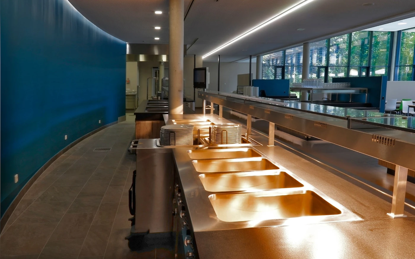 Eine moderne, leere Cafeteria mit einer langen Edelstahltheke, Speisenwärmern, gestapelten Tellern, einer blauen Wand auf der linken Seite und großen Fenstern, durch die Tageslicht hereinströmt – ein einladender Raum, der von SCHATZ Architektur sorgfältig gestaltet wurde, um innovative Projektentwicklung widerzuspiegeln.