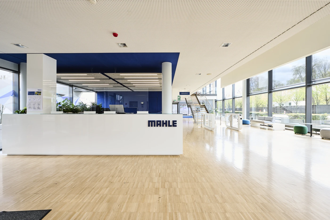 Projekte Moderne Büro-Lobby mit einem weißen Empfangstresen mit der Aufschrift „MAHLE“, hellem Holzfußboden, großen Fenstern, Topfpflanzen und einer Treppe, die in ein oberes Stockwerk führt. Der Raum ist hell und offen und wird von Tageslicht durchflutet, was den durchdachten Entwurfsprozess widerspiegelt, der jedes Detail dieses Standorts in Schorndorf über alle Leistungsphasen hinweg geprägt hat.