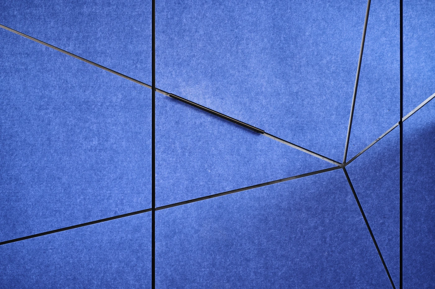 Eine abstrakte blaue Wand mit sich kreuzenden schwarzen Linien, die geometrische Formen wie Dreiecke und Vierecke bilden – was an Elemente aus den Bereichen „Entwurf“ und „Projektentwicklung“ erinnert. Die Oberfläche wirkt strukturiert, und in der Mitte befindet sich ein einzelner horizontaler Balken, der an die präzise „Generalplanung“ im modernen Design erinnert.