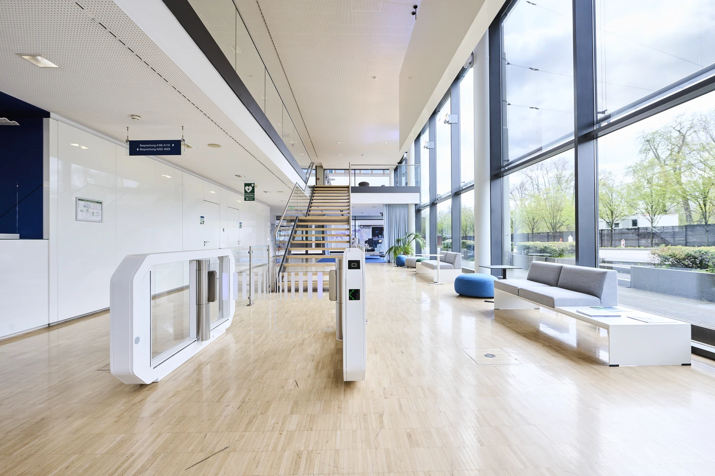 Projekte Moderne Büro-Lobby mit Holzboden, großen Fenstern, Sitzecken, einem Sicherheitstor und einer zentralen Treppe. Der Raum wird von Tageslicht durchflutet, und draußen sind Bäume zu sehen – ein Beispiel für die durchdachte Generalplanung, die jedes Detail in allen Leistungsphasen des Bauvorhabens in Schorndorf geprägt hat.