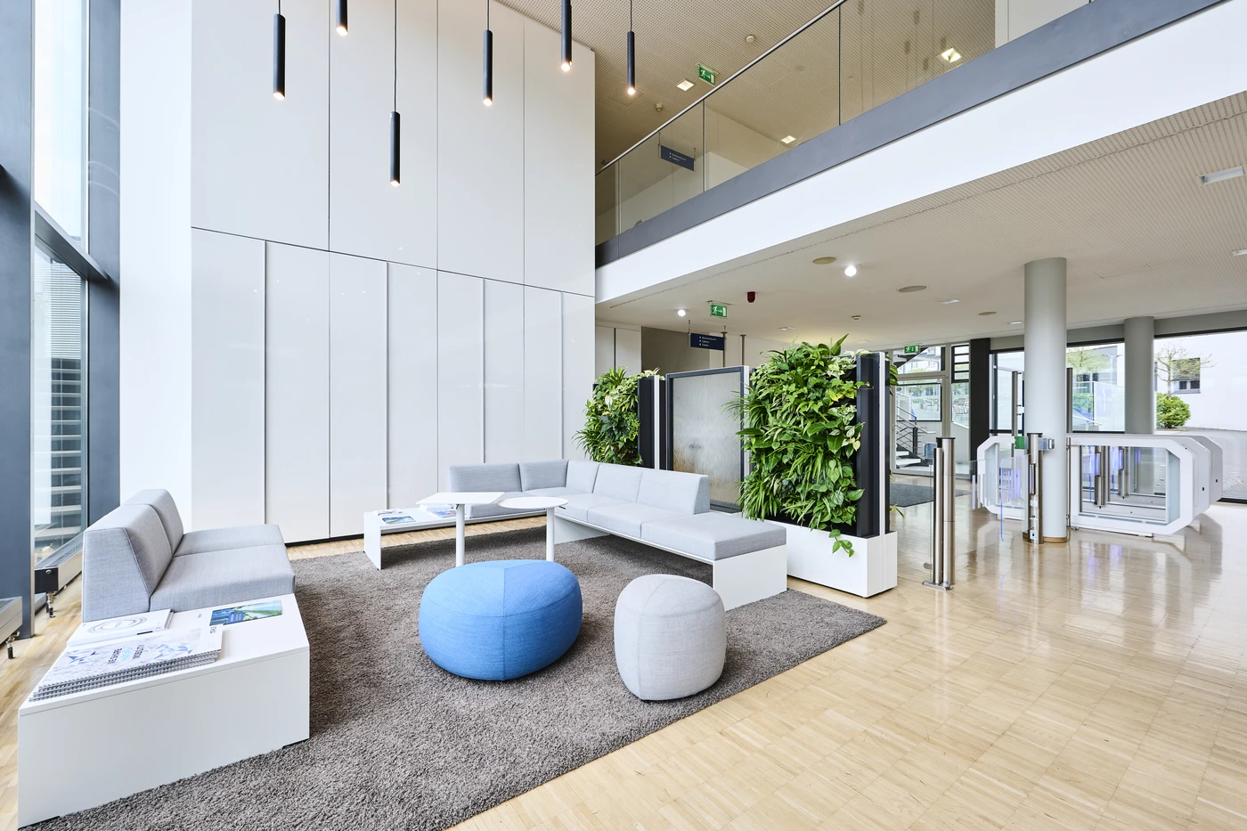 Projekte Moderne Büro-Lobby mit grauen Sofas, blauen und grauen Sitzkissen, weißen Tischen, Zimmerpflanzen und einem braunen Teppich auf hellem Holzboden. Große Fenster und hohe Decken sorgen für natürliches Licht in diesem durchdachten Raum in Schorndorf – ein hervorragendes Beispiel für moderne Generalplanung, die Form und Funktion nahtlos miteinander verbindet.