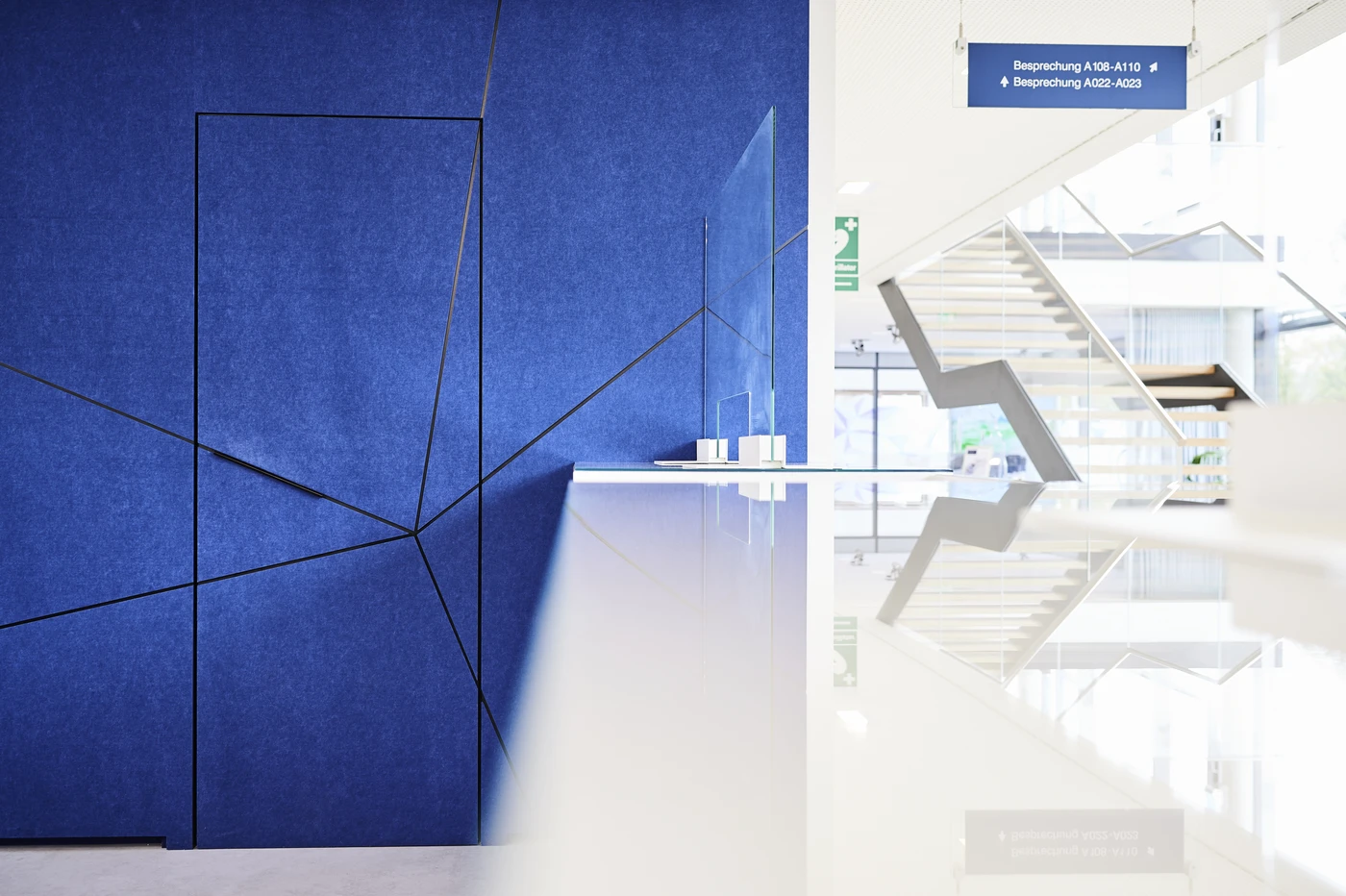 Projekte Modernes Interieur mit einer geometrischen blauen Wand auf der linken Seite, einer weißen, spiegelnden Theke, einer Treppe im Hintergrund und einem blauen Schild mit weißer Beschriftung, das von der Decke hängt und auf Besprechungsräume hinweist – ein inspirierender Raum, der Form und Funktion für jede Entwurfs- oder Projektentwicklungsphase nahtlos miteinander verbindet.