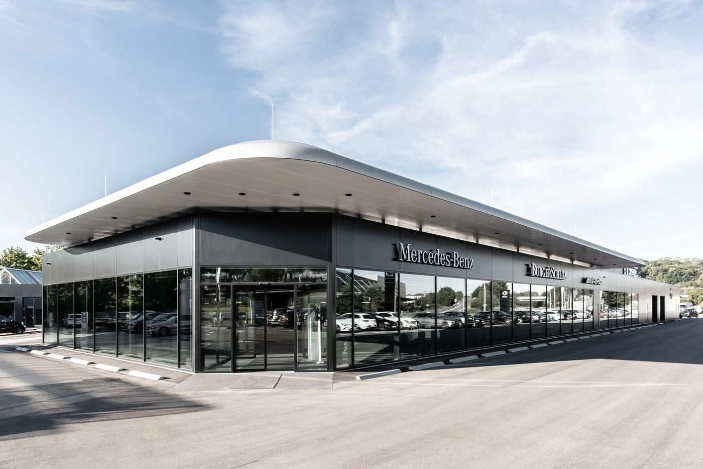 Projekte Modernes Mercedes-Benz-Autohaus mit großen Glasfenstern, metallischer Fassade und durch das Glas sichtbaren geparkten Autos. Der Entwurf in Schorndorf besticht durch klare architektonische Linien und Transparenz und fügt sich nahtlos in den blauen Himmel und das umgebende Grün im Hintergrund ein.