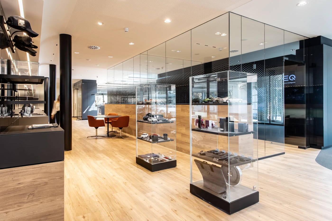Projekte Moderner Showroom in Schorndorf mit Glasvitrinen, in denen verschiedene Produkte ausgestellt sind, Holzfußboden, Regalen mit Artikeln auf der linken Seite und einer kleinen Sitzecke mit braunen Stühlen im Hintergrund. Der Raum ist hell und geräumig und zeugt von einer durchdachten Gesamtplanung und Projektentwicklung.