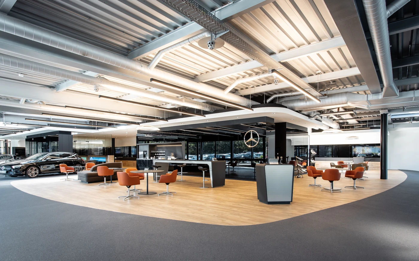 Projekte Moderner Showroom eines Autohauses mit eleganten Möbeln, hellem Holz und dunklem Bodenbelag, mehreren ausgestellten Fahrzeugen, orangefarbenen Stühlen und einem großen Mercedes-Benz-Logo an der Wand. Der Entwurf hebt die Verwendung von freiliegenden Leitungen und industrieller Beleuchtung an der Decke hervor und spiegelt damit einen zeitgemäßen Ansatz bei der Projektentwicklung in Schorndorf wider.