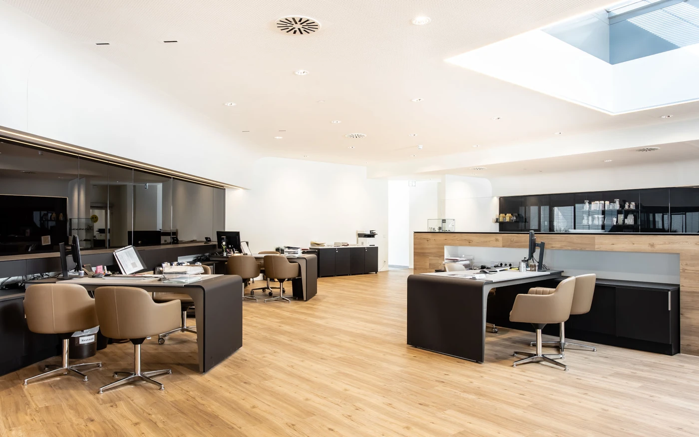Projekte Ein modernes Büro mit mehreren freien Schreibtischen und beigen Stühlen auf einem Holzboden, heller Beleuchtung, weißen Wänden und einem großen Oberlicht. Im gesamten Arbeitsbereich sind Computer, Büromaterial und Regale zu sehen – eine professionelle Umgebung, die von SCHATZ Architektur im Rahmen eines Generalplanungsprojekts in Schorndorf sorgfältig gestaltet wurde.