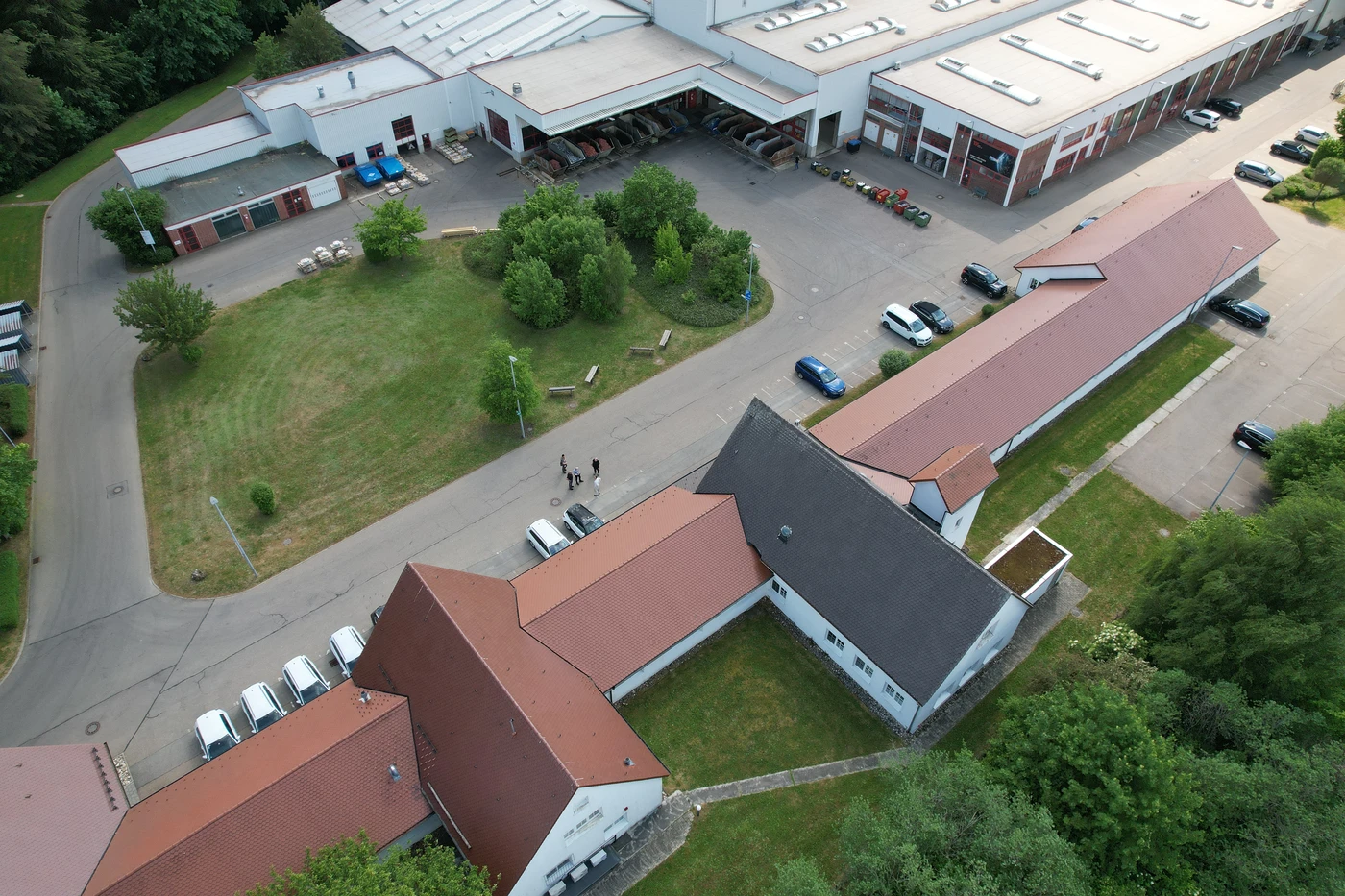 Luftaufnahme eines großen Industriegebäudes mit roten Dächern, geparkten Autos, vereinzelten Bäumen und Menschen, die auf gepflasterten Wegen in der Nähe von grünen Rasenflächen spazieren gehen – ein eindrucksvolles Beispiel für die Generalplanung in Schorndorf, das eine durchdachte Projektentwicklung verdeutlicht.