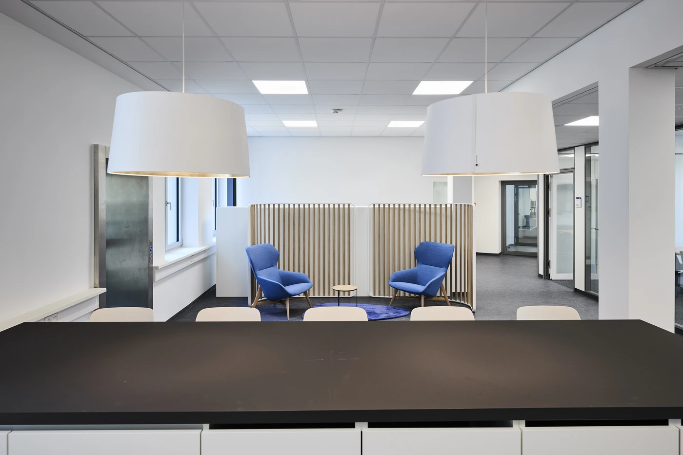 Projekte Ein von SCHATZ Architektur entworfener moderner Büroraum mit einem schwarzen Tisch, weißen Stühlen und zwei großen weißen Pendelleuchten. Im Hintergrund unterstreicht eine Sitzecke mit zwei blauen Sesseln und einem kleinen runden Tisch auf einem blauen Teppich die durchdachte Projektentwicklung und Generalplanung, die dieses anspruchsvolle Ambiente auszeichnen.