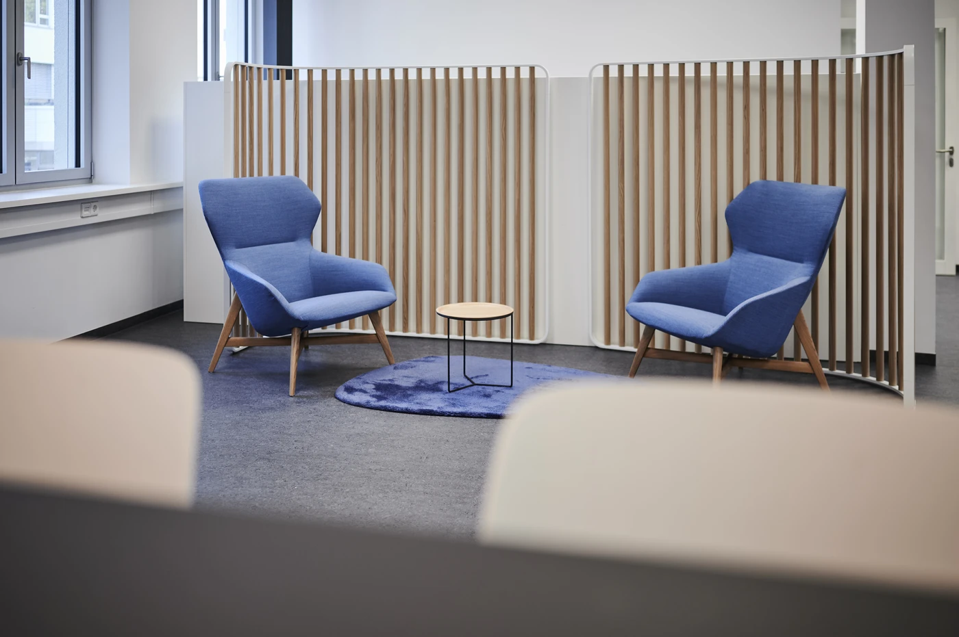 Projekte Zwei moderne blaue Sessel stehen sich gegenüber, dazwischen steht ein kleiner runder Holztisch auf einem blauen Teppich – in einem Büroraum mit einem Raumteiler aus Holzlamellen: eine einladende Kulisse, die den durchdachten Ansatz der Generalplanung und die detaillierte Koordination widerspiegelt, wie sie oft in Schorndorfs jüngsten Leistungsphasen zu beobachten sind.