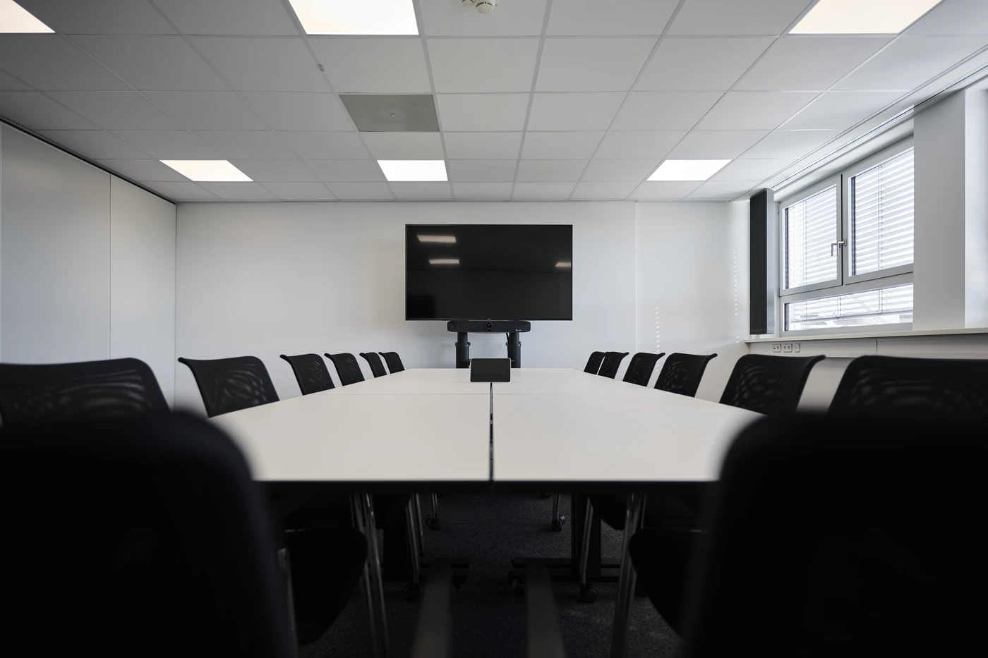 Projekte Ein moderner, leerer Konferenzraum mit einer großen Leinwand an der Vorderseite, weißen Tischen, die in U-Form angeordnet sind, und schwarzen Stühlen auf beiden Seiten. Große Fenster auf der rechten Seite lassen Tageslicht herein – ideal für Besprechungen rund um Entwurf und Projektentwicklung in allen Leistungsphasen.