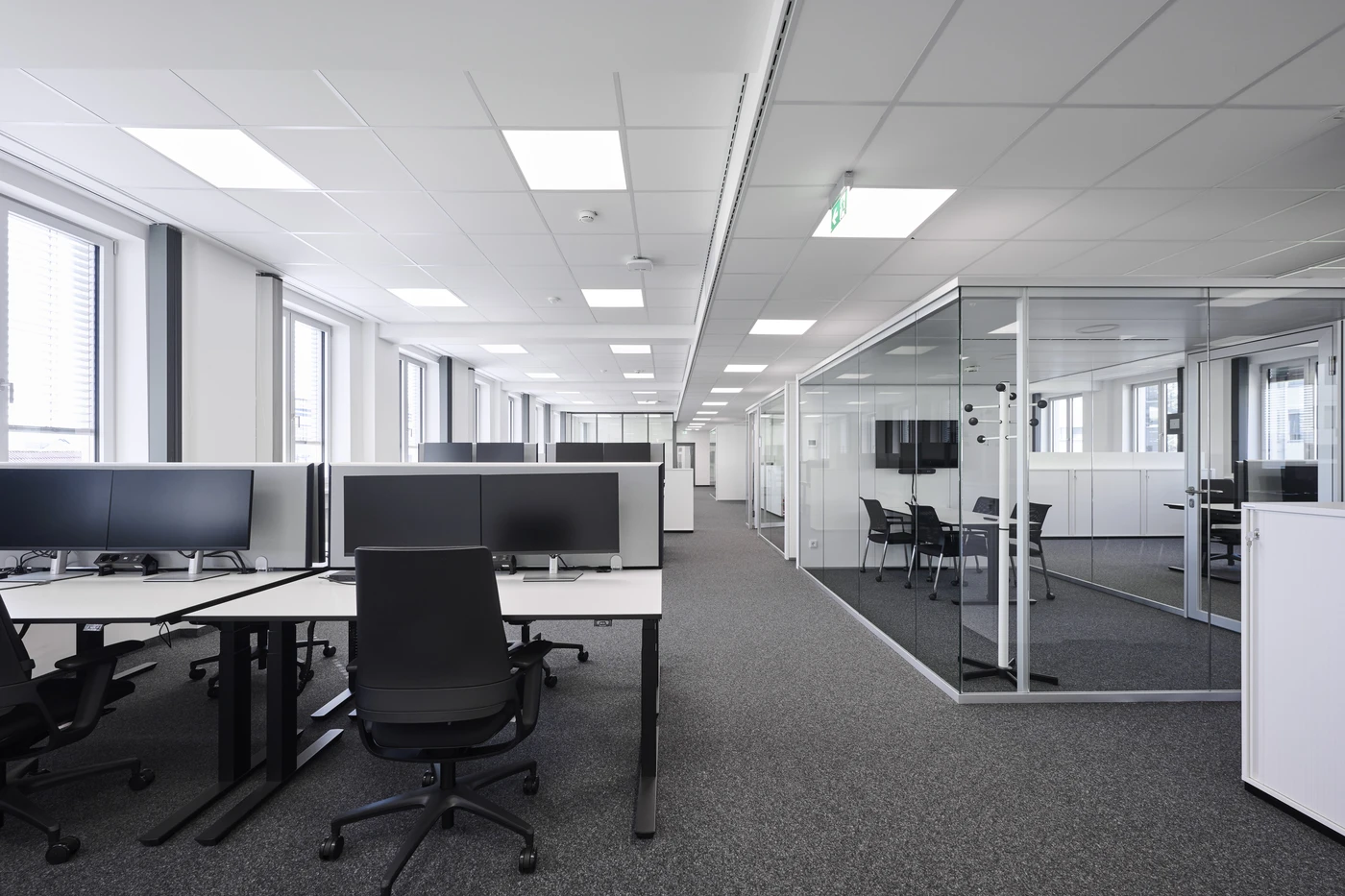 Projekte Ein modernes Büro mit Reihen leerer Schreibtische, Computermonitoren, ergonomischen Stühlen und einem verglasten Besprechungsraum auf der rechten Seite. Große Fenster sorgen für Tageslicht und schaffen einen aufgeräumten und übersichtlichen Arbeitsbereich – ein beeindruckender Entwurf von SCHATZ Architektur, der die zeitgenössische Designphilosophie in Schorndorf widerspiegelt.