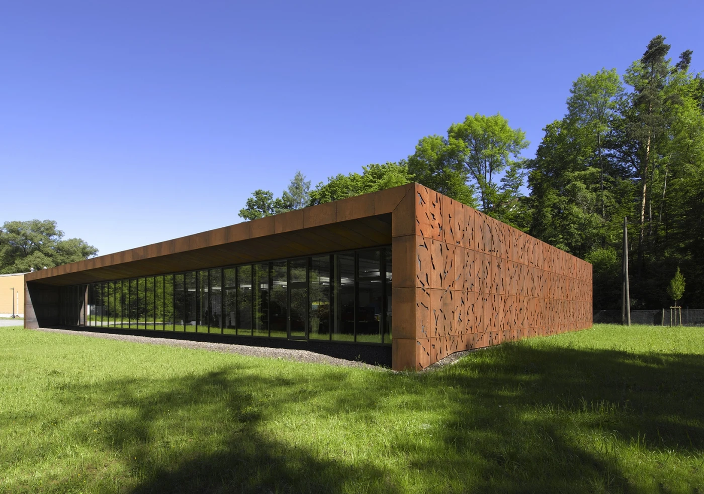 Ein modernes, niedriges, rechteckiges Gebäude mit rostfarbenen, strukturierten Metallwänden und großen Glasfenstern, entworfen von SCHATZ Architektur, das auf einer Rasenfläche mit Bäumen steht und vor dem Hintergrund eines strahlend blauen Himmels – ein vorbildliches Projekt im Bereich innovativer Projektentwicklung für Schorndorf.