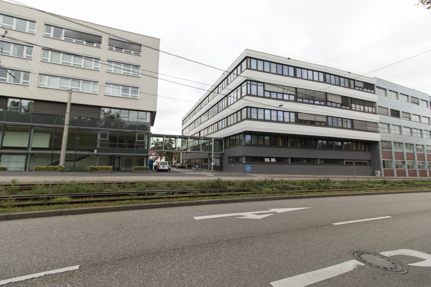 Moderne Bürogebäude mit großen Fenstern sind durch eine Glasbrücke miteinander verbunden, was von einem durchdachten Entwurf zeugt. In der Nähe des Eingangs steht ein Auto, und im Vordergrund verlaufen Straßenbahnschienen entlang der Straße. Die Szene, die in Schorndorf unter bewölktem Himmel spielt, verdeutlicht verschiedene Leistungsphasen der Stadtentwicklung.