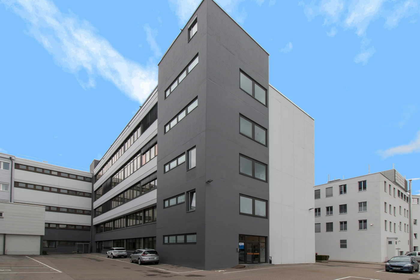 Dieses Bild zeigt ein modernes, mehrstöckiges Bürogebäude mit grauen und weißen Außenwänden, großen Fenstern und mehreren vor dem Gebäude geparkten Autos unter strahlend blauem Himmel und verdeutlicht damit die Präzision von Generalplanung in allen Leistungsphasen eines Projekts in Schorndorf.