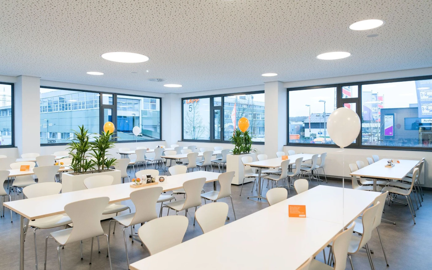 Eine helle, moderne Cafeteria mit weißen Tischen und Stühlen, großen Fenstern, Grünpflanzen und einigen orange-weißen Luftballons. Der Raum, der von einer durchdachten Planung und Projektentwicklung zeugt, wirkt klar und leer, und von hier aus sind die Gebäude der Stadt zu sehen.